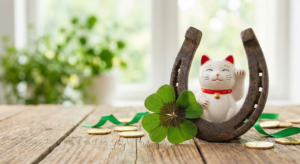A collection of lucky symbols, including a white Maneki Neko beckoning cat nestled inside a rustic horseshoe, a green four-leaf clover, and scattered gold coins, all arranged on a wooden table by a bright window.