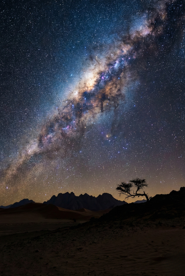 Milky Way galaxy arching over a dark silhouette desert mountain landscape.