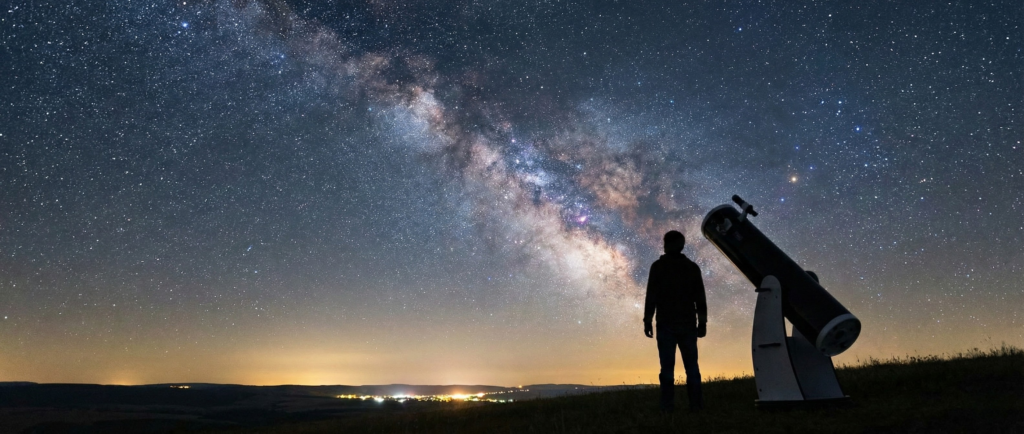 A silhouette of a person standing next to a telescope under the Milky Way, contemplating the cosmic origins found in their Starseed birth chart.